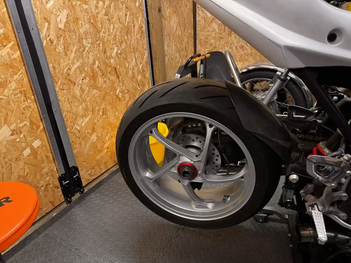 A close-up photo of a motorcycle's rear wheel, showing a silver rim, black tire, and yellow brake caliper, parked indoors on…