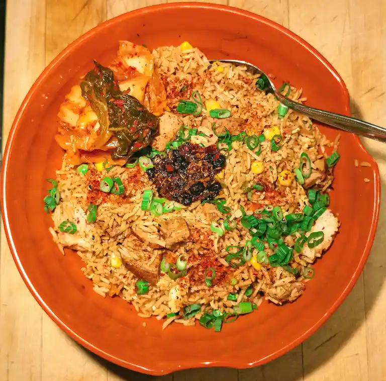 An overhead photo of a vibrant orange bowl filled with seasoned fried rice, topped with chopped green onions, corn, meat…