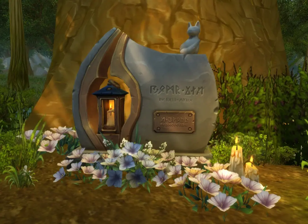 A gravestone shaped like a book with a cat figurine on top, a lantern inside, and flowers around its base. The stone has…
