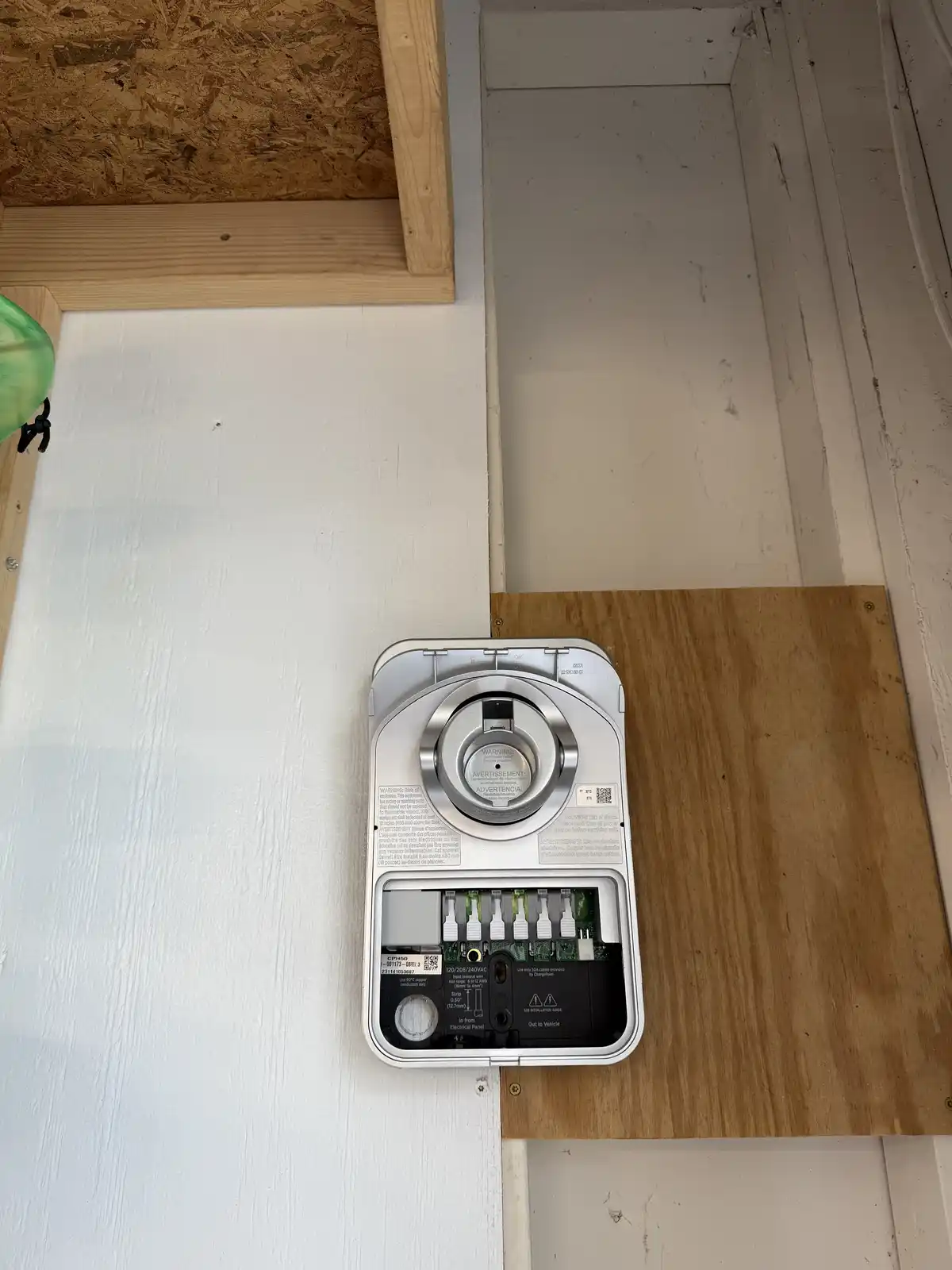 A silver electric vehicle charging station is mounted on a white wall next to a wooden board, with its back panel open…