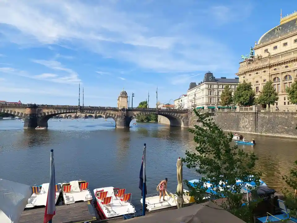 A scenic photo of a river with a large stone bridge and ornate buildings under a blue sky. Several paddle boats are docked…