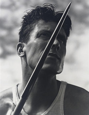 A black-and-white photo of a man in a tank top holding a javelin, looking upward with a serious expression. The text…