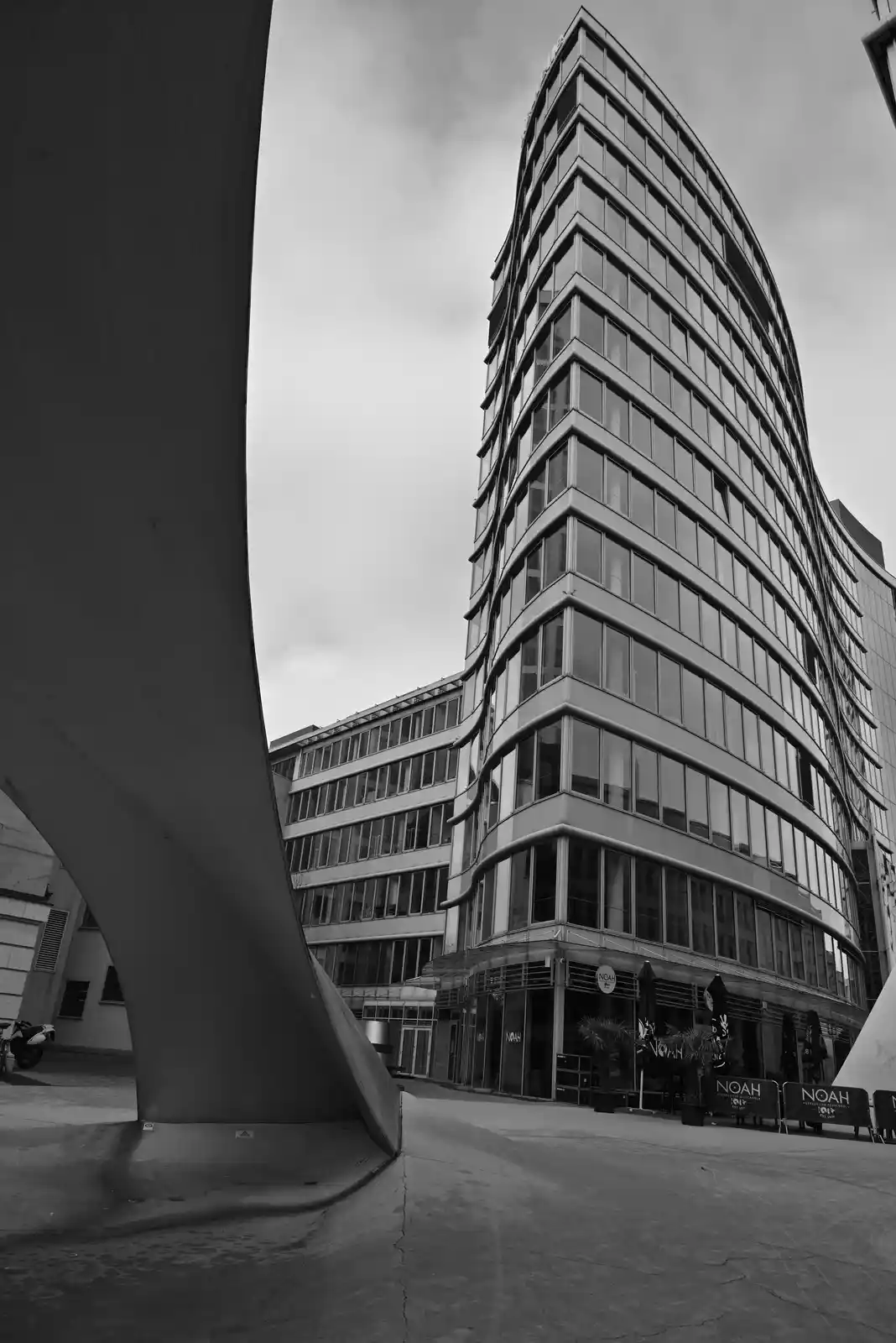 A black-and-white photo shows a modern, curved glass skyscraper viewed from a low angle, framed by a large, curved…
