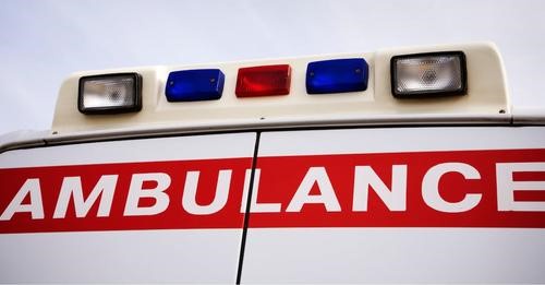 A close-up photo of the top of a white ambulance, showing its red and blue emergency lights and the word 'AMBULANCE' in…