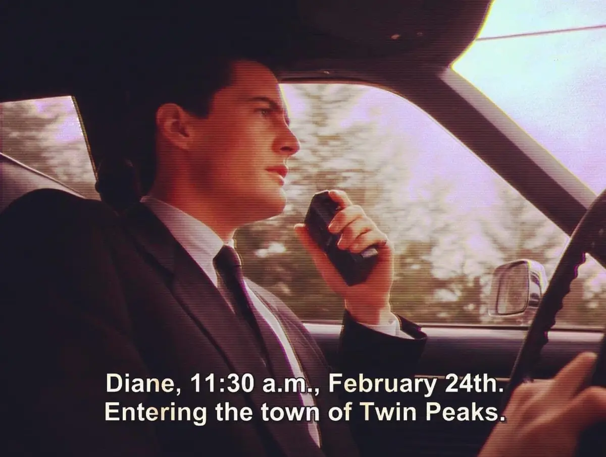A man in a suit and tie sits in the driver's seat of a car, holding a walkie-talkie to his ear while looking forward. The…