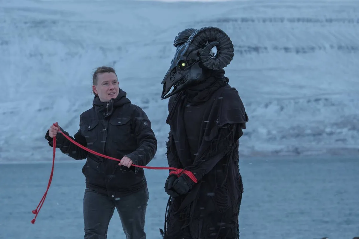 A person in a black winter jacket holds a red leash attached to a tall, costumed figure wearing a large, menacing ram mask…