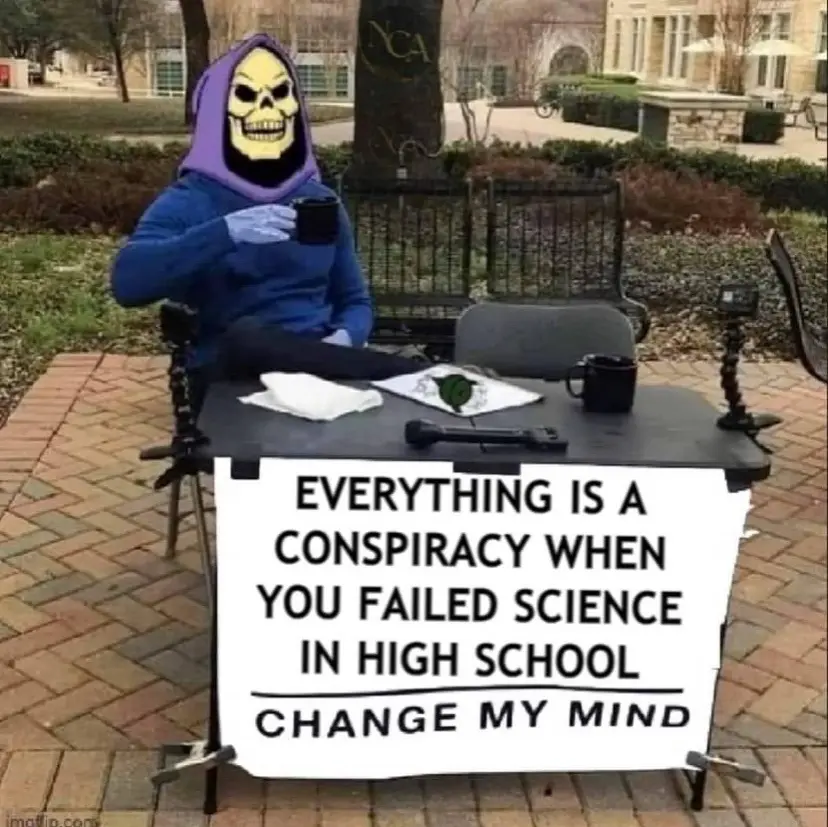 A person wearing a Skeletor mask and blue hoodie sits at an outdoor table with a mug, surrounded by a microphone, camera…