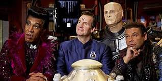 Four characters from the TV show 'Doctor Who' are shown seated around a table with a large golden object. The character on…