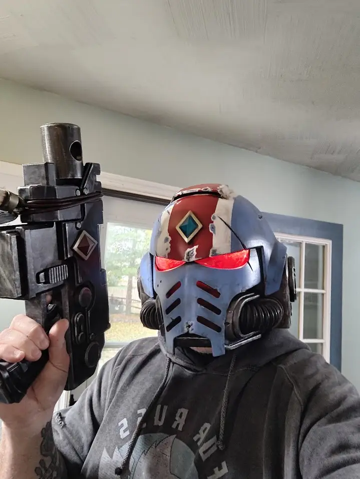 A person wearing a heavily weathered, red-and-blue armored helmet with glowing red eyes and holding a large, detailed prop…