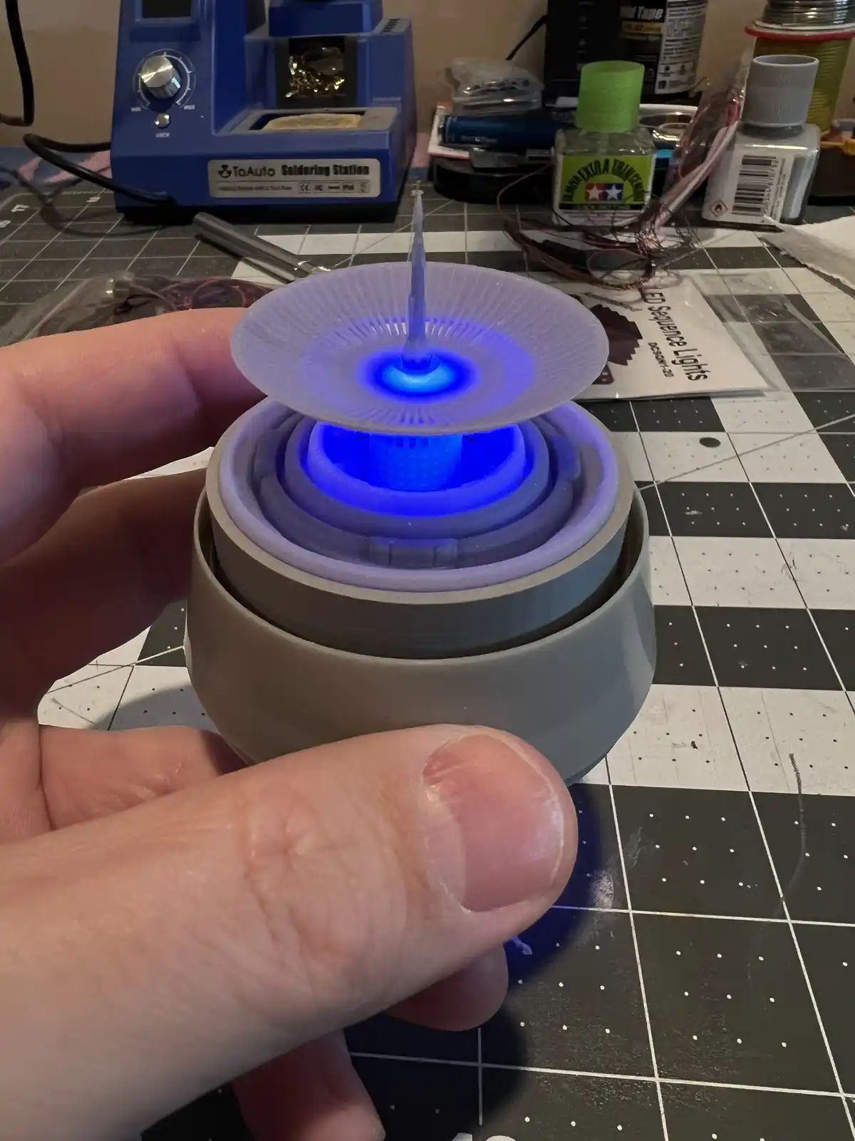 A hand holds a 3D-printed object resembling a lightsaber hilt, with a glowing blue blade effect emanating from its center…