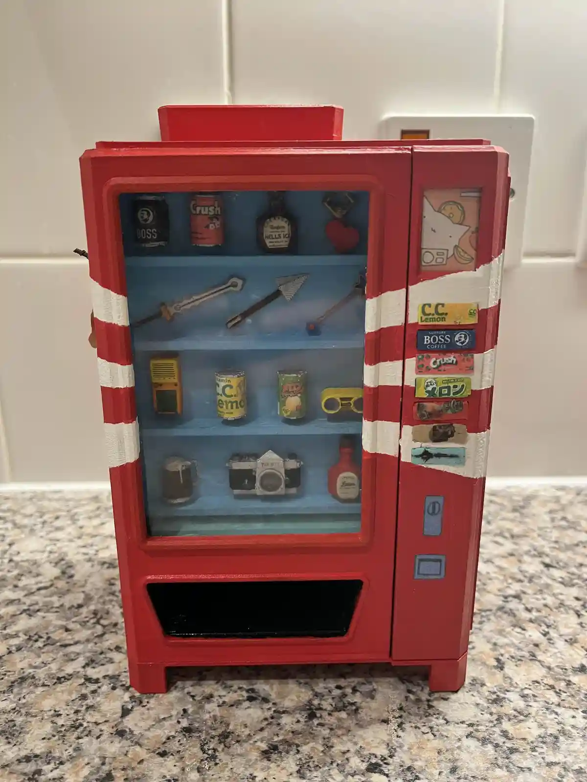 A red and white striped miniature vending machine sits on a granite countertop, its glass front revealing shelves filled…
