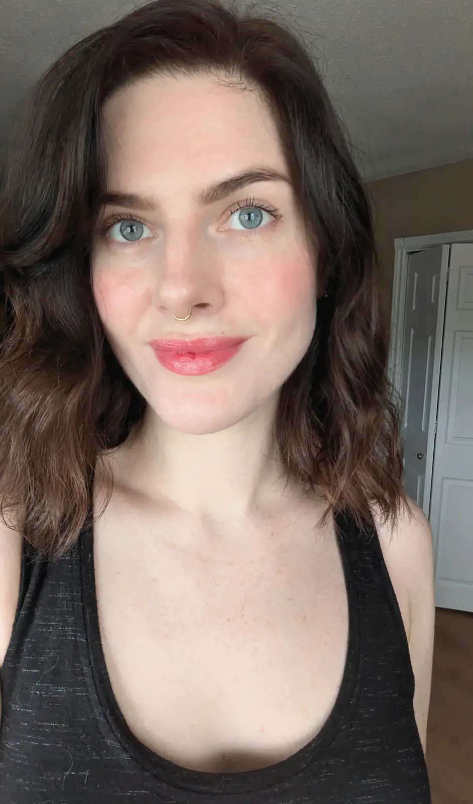 A selfie of a woman with brown wavy hair, blue eyes, and a gold nose ring, wearing a black tank top. She has a slight smile…