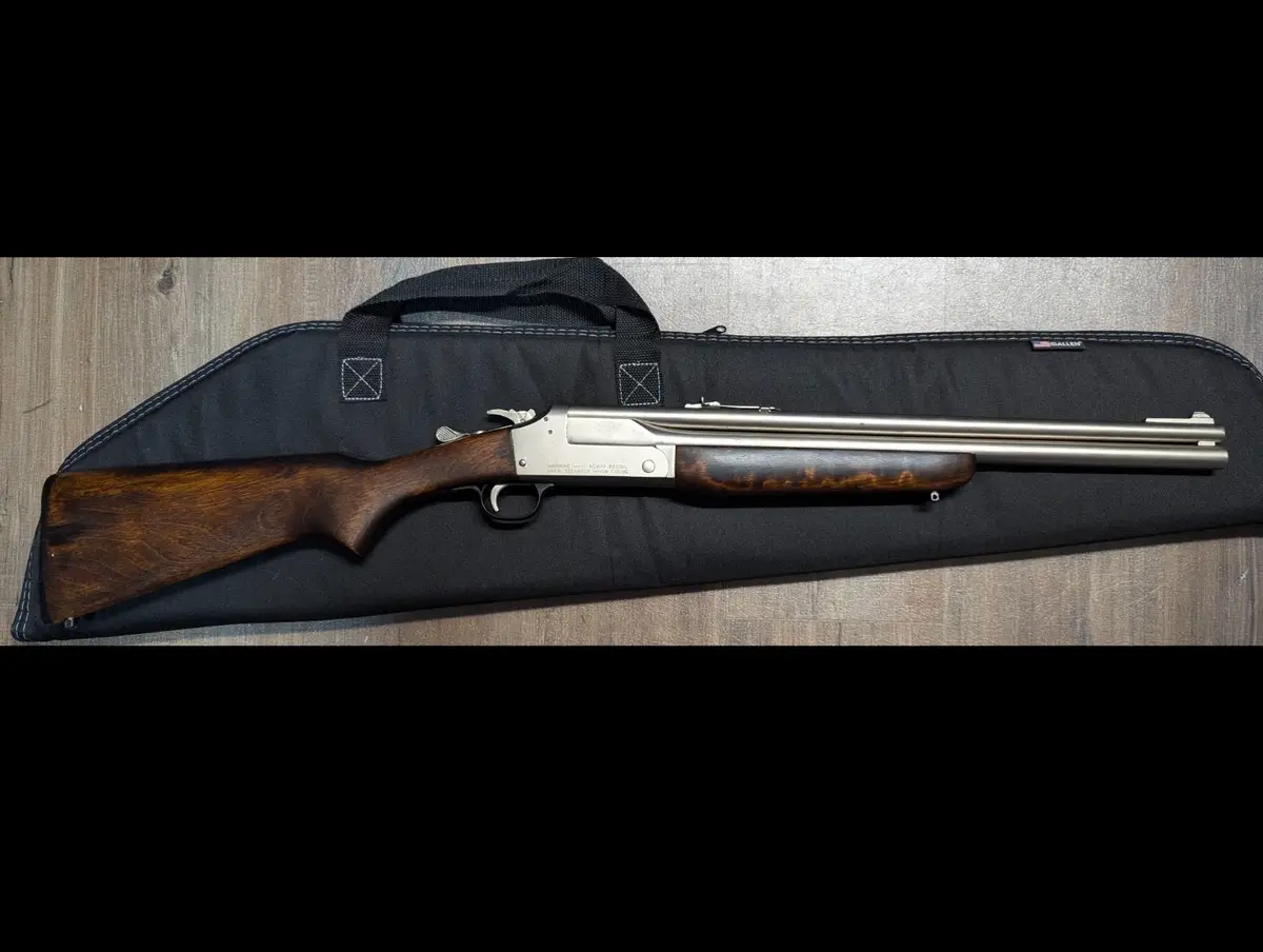 A double-barreled shotgun with a wooden stock and metal barrel rests horizontally on a black fabric gun case, which is…