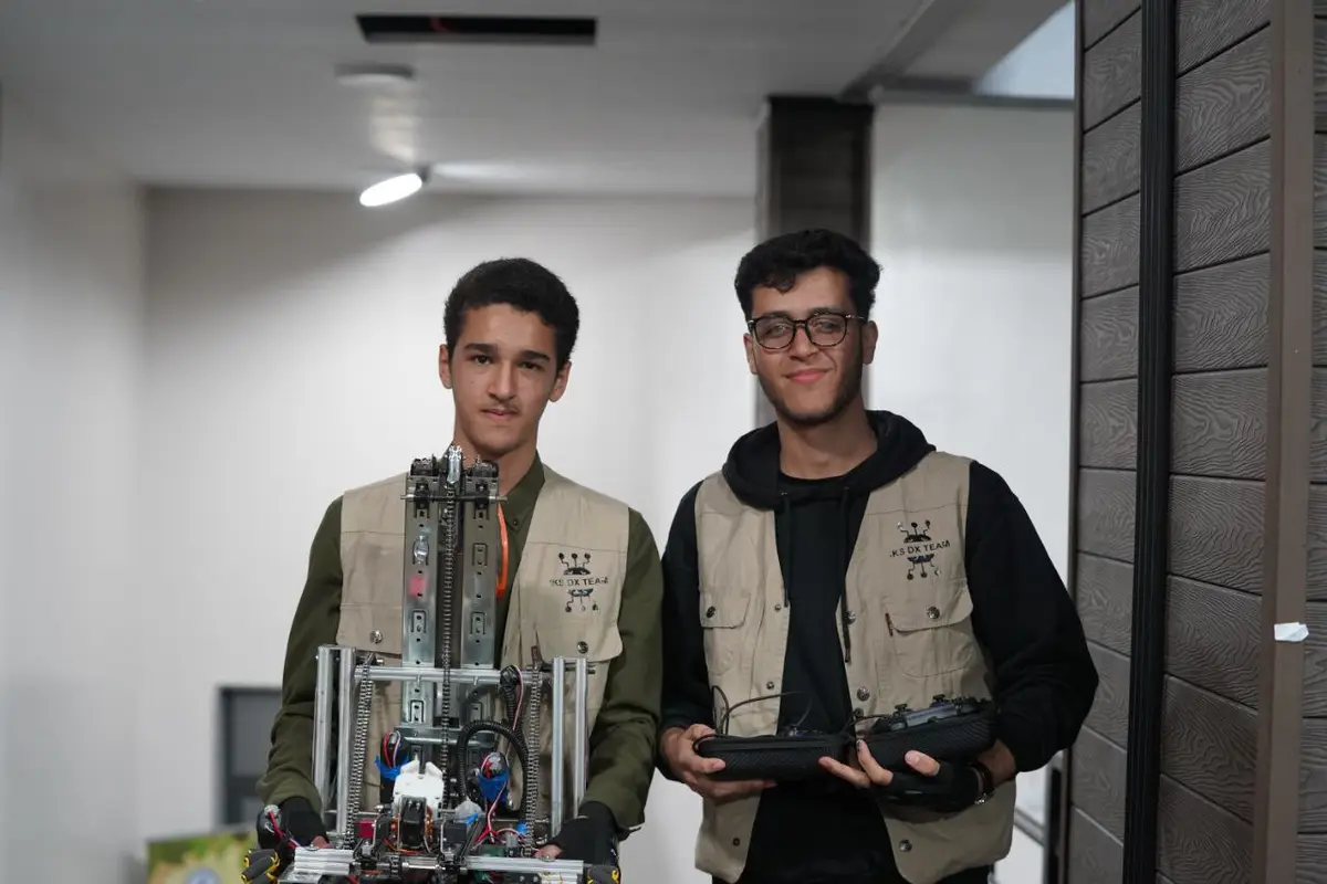 Two young men wearing matching beige vests with 'J.K.S DX TEAM' logos stand indoors. The man on the left holds a complex…