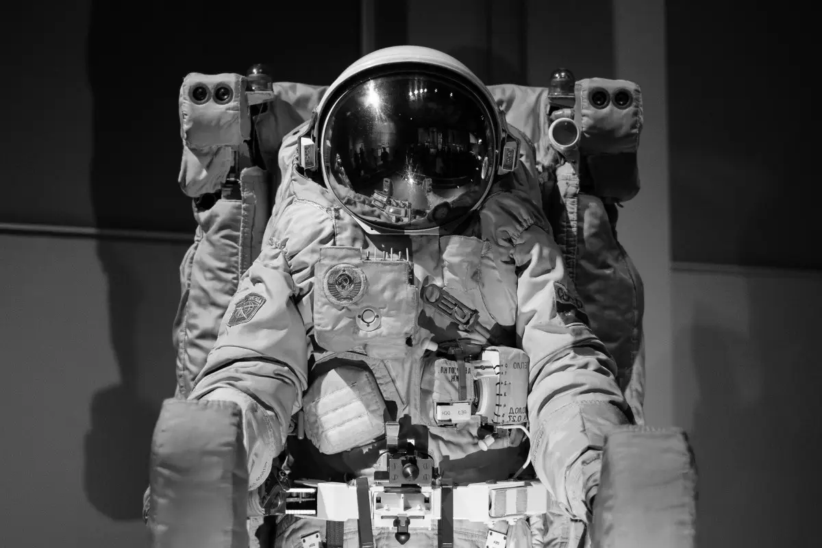 A black-and-white photo shows a person in a full Soviet-era space suit seated in a chair, with the reflective helmet…