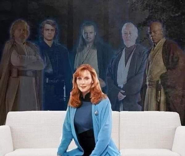 A woman with red hair wearing a blue jacket sits on a white couch, looking directly at the camera. Behind her, a faded image…
