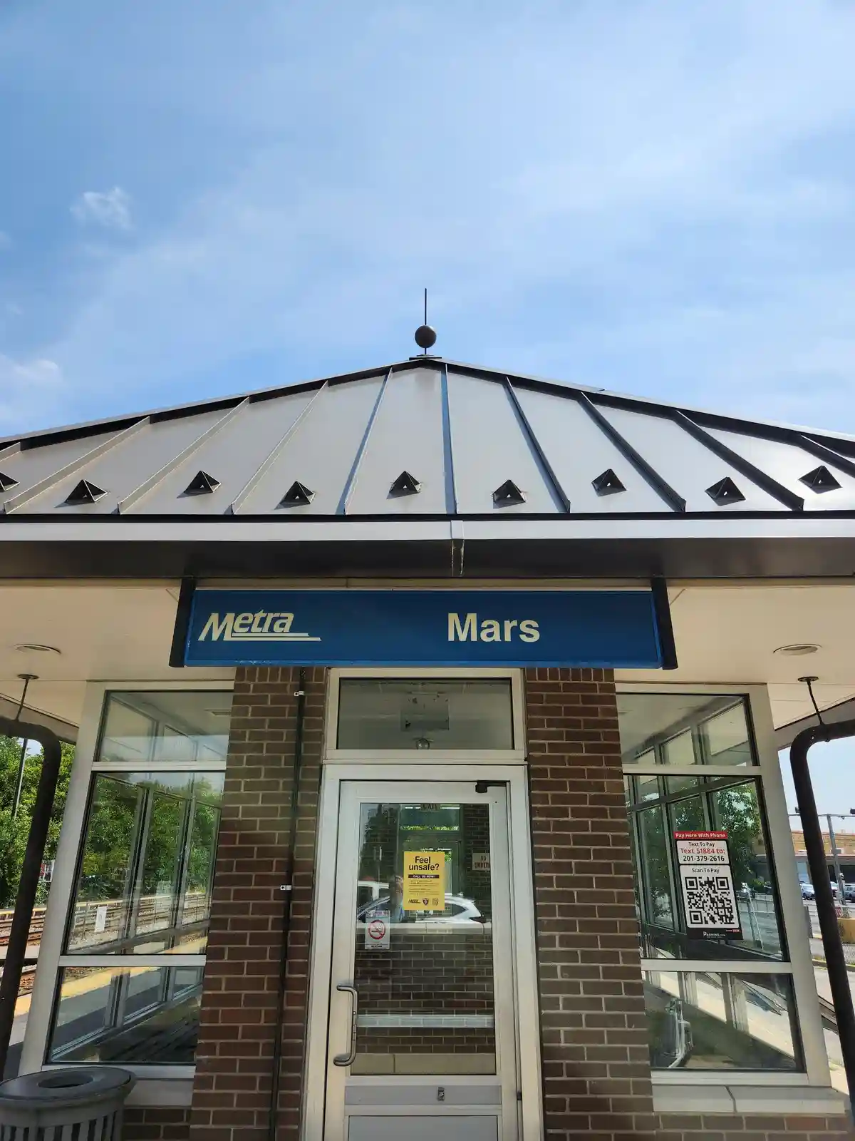 A photo of a Metra train station entrance with a blue sign that reads 'Metra Mars' under a white and black peaked roof. The…