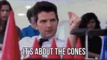 A man in a white shirt and bowtie sits in an audience surrounded by red traffic cones, repeatedly saying 'IT'S ABOUT THE…