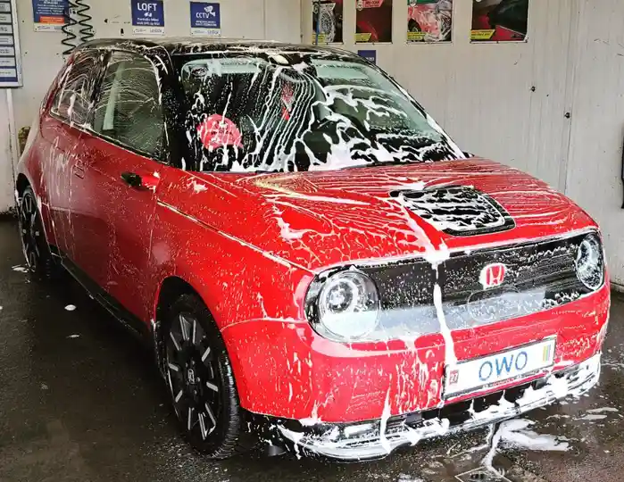 A red Honda car is heavily covered in white soap suds during a wash, with foam dripping from its body and windows. The…