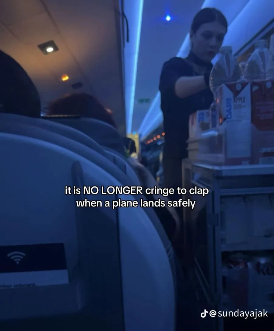 A blurry photo taken from inside an airplane cabin shows a flight attendant standing by a cart with drinks, including Oasis…