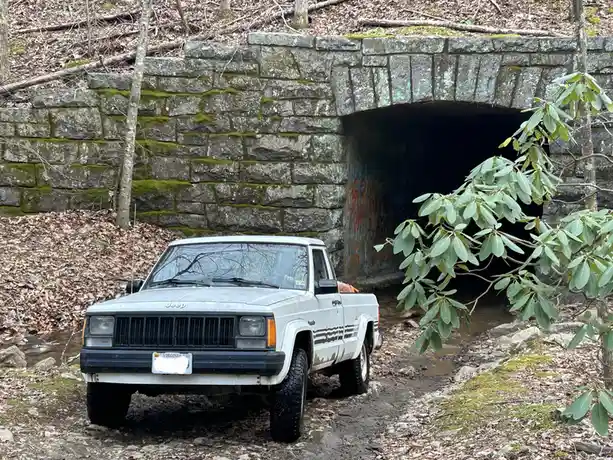 A white, older model Jeep Cherokee pickup truck is parked on a muddy, leaf-covered trail in front of a stone archway tunnel…