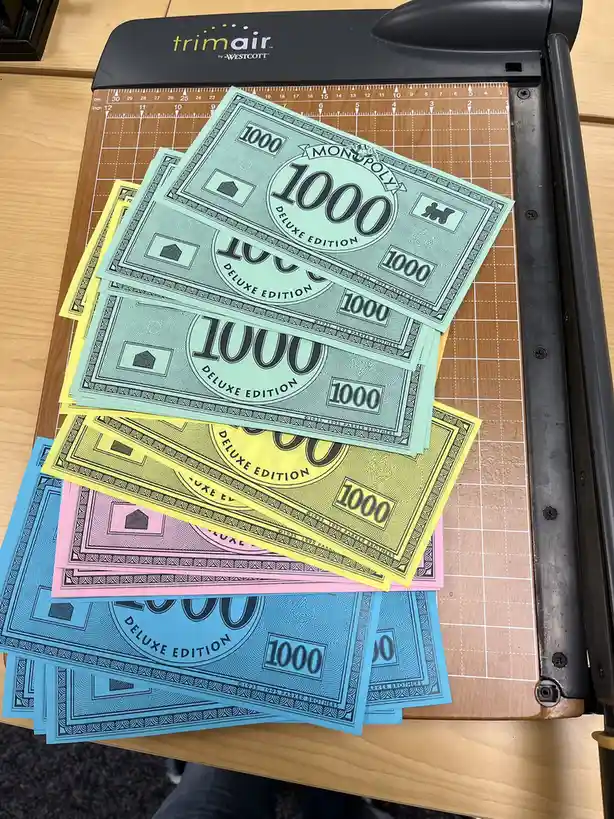 Stacks of colorful Monopoly Deluxe Edition $1000 bills are piled on a cutting mat next to a black Trimair paper trimmer. The…