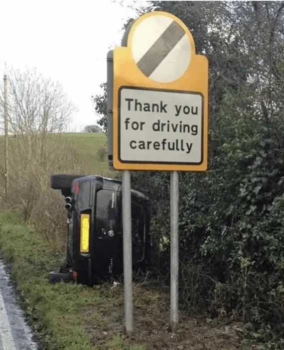 A black car is overturned on its side in a grassy ditch next to a road, partially obscured by bushes. A yellow road sign…
