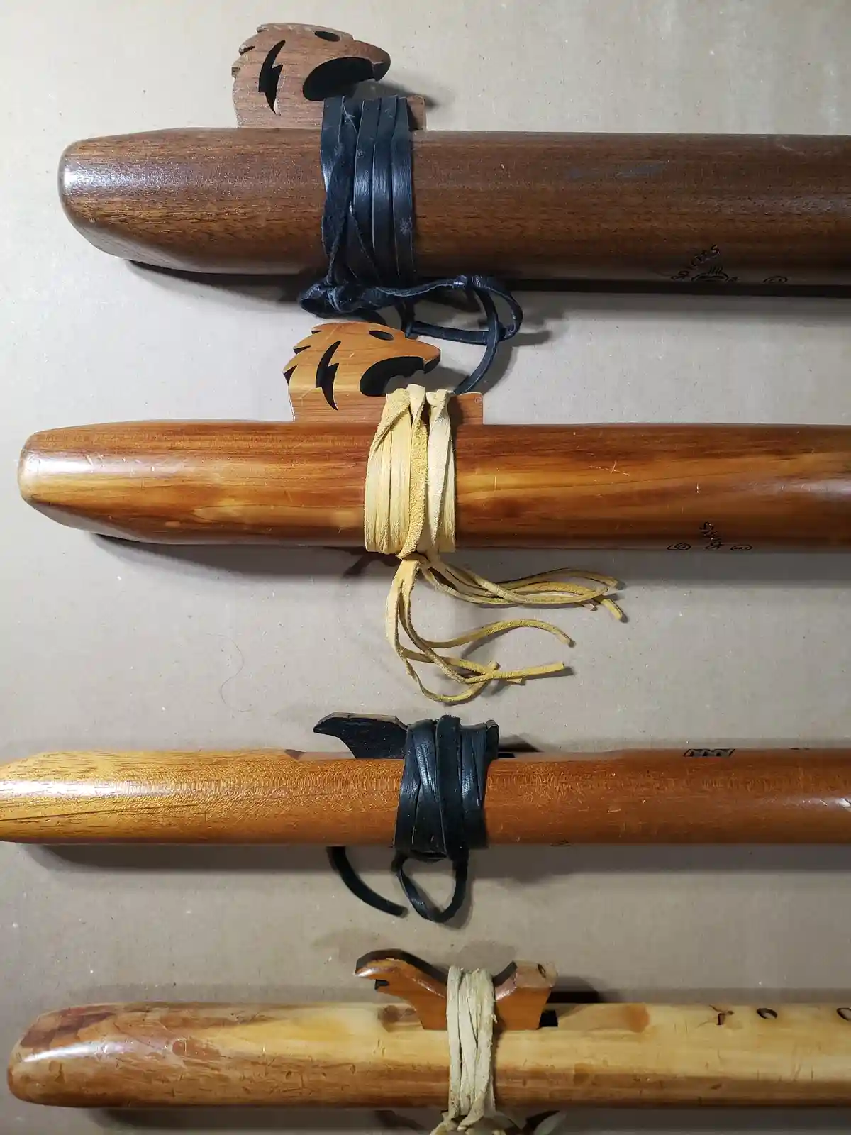 Four wooden flutes with decorative eagle-head mouthpieces and leather or suede straps are arranged vertically on a plain…
