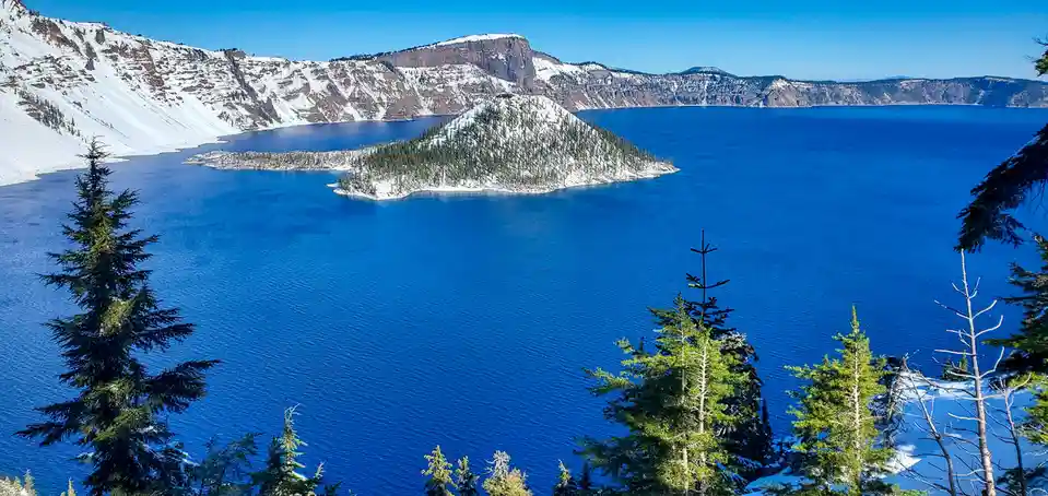 A scenic winter photograph of Crater Lake, featuring deep blue water, snow-covered volcanic rim, and a forested island…