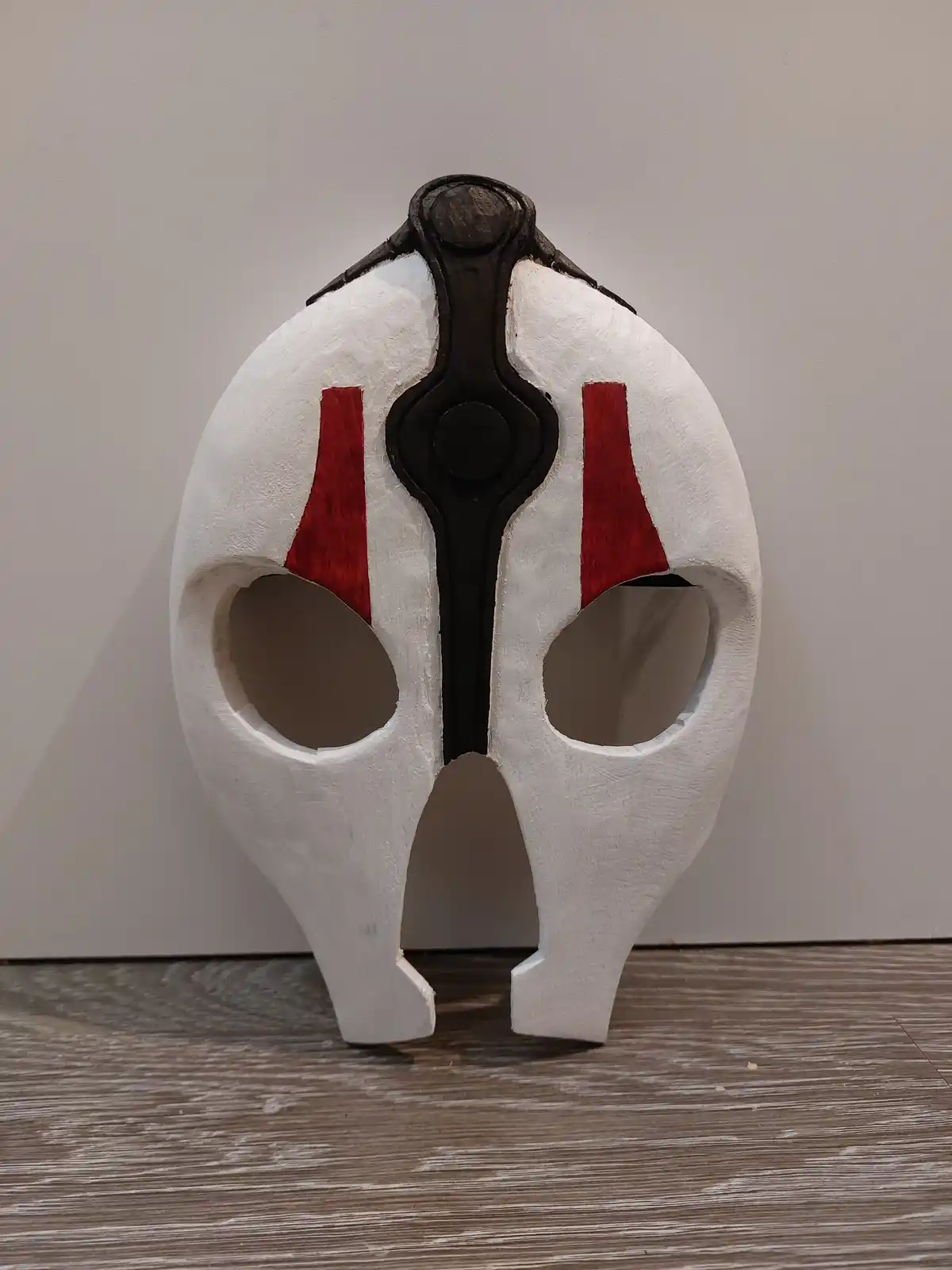 A handmade white cosplay mask with red triangular markings on the cheeks and a black central design, standing upright on a…