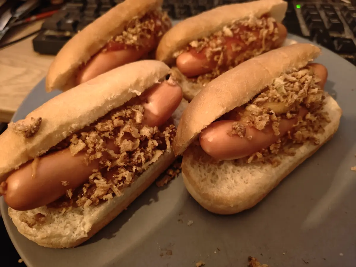 A close-up photo shows four hot dogs in buns on a gray plate, each topped with a generous amount of crispy fried onions and…