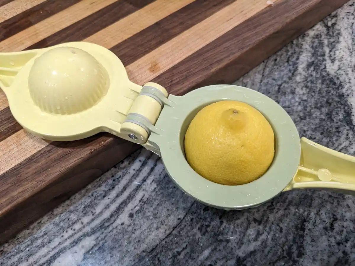 A yellow and light green citrus juicer sits on a wooden cutting board and granite countertop, with a whole lemon placed in…