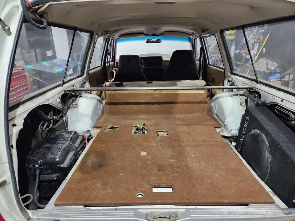 The image shows the interior of a white station wagon with its rear seats removed, revealing a brown carpeted floor with…