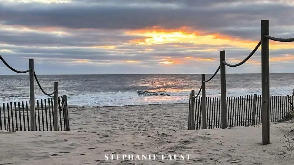 A serene beach scene at sunset with waves crashing onto the shore, framed by wooden fences and rope posts. The sky is…