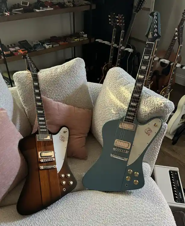 Two electric guitars, one sunburst and one teal, are propped up on a white textured couch with pillows. In the background…