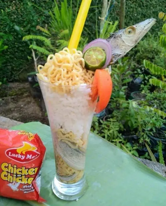 A tall glass contains layered instant noodles, rice, and a whole fish head, garnished with lime, onion, and tomato slices…