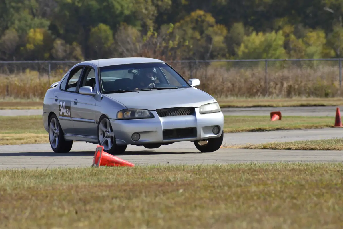 A silver sedan with the number 34XSA on its door is driving on a paved track, narrowly missing an orange traffic cone that…