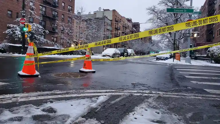 A snowy urban street intersection is blocked off by yellow 'FIRE LINE: DO NOT CROSS' caution tape strung between orange…