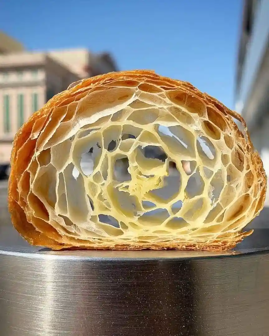 A close-up photo shows a perfectly sliced croissant revealing its intricate, honeycomb-like flaky interior with golden-brown…