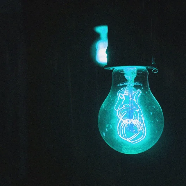 A glowing light bulb with a blue-green hue hangs against a dark background, its filament clearly visible inside the glass…