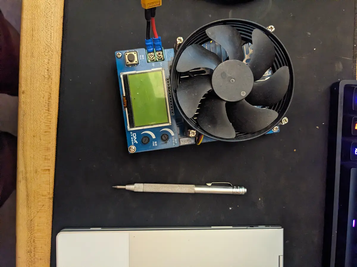 A blue DIY electronics board with a small green screen and a black computer fan mounted on it sits on a black desk next to a…
