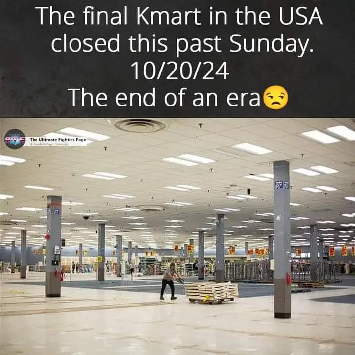 An empty, vast Kmart store interior with fluorescent lighting and pillars, a lone person pushing a cart of wooden pallets…