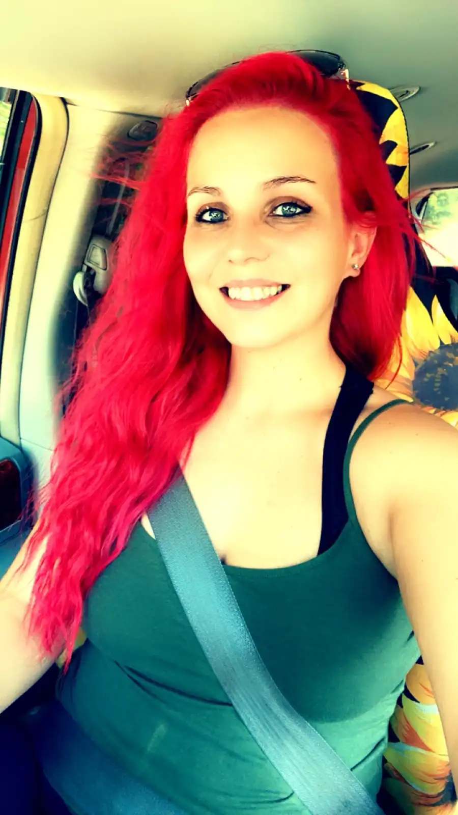 A woman with vibrant red hair is taking a selfie inside a car, wearing a green tank top and a seatbelt, with a…