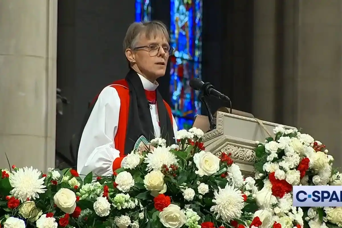 A woman in clerical attire, including a white shirt, red stole, and black scarf, stands at a decorated pulpit in a church…
