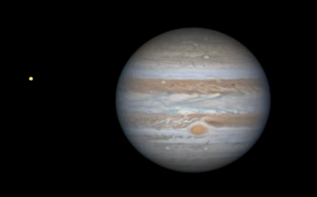 A space photograph shows the planet Jupiter with its prominent striped cloud bands and the Great Red Spot, with a small…