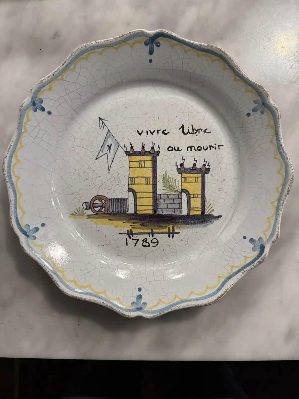 A ceramic plate with a scalloped edge and crackled glaze features a hand-painted scene of two yellow towers with cannons, a…