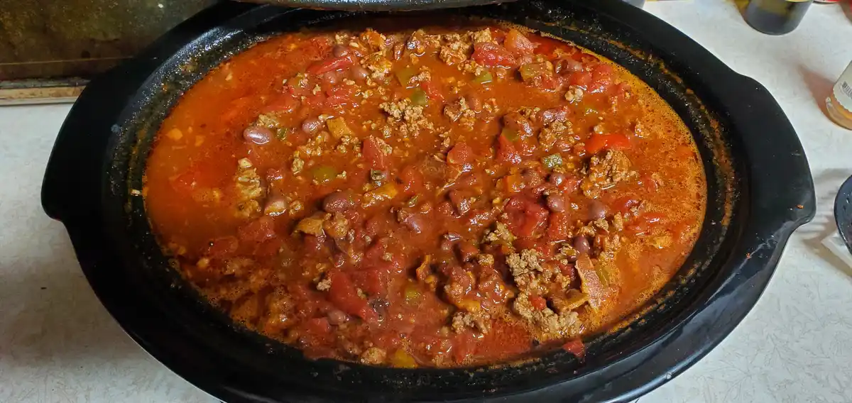 A black oval slow cooker is filled with a reddish-orange stew containing ground meat, beans, diced tomatoes, and green…