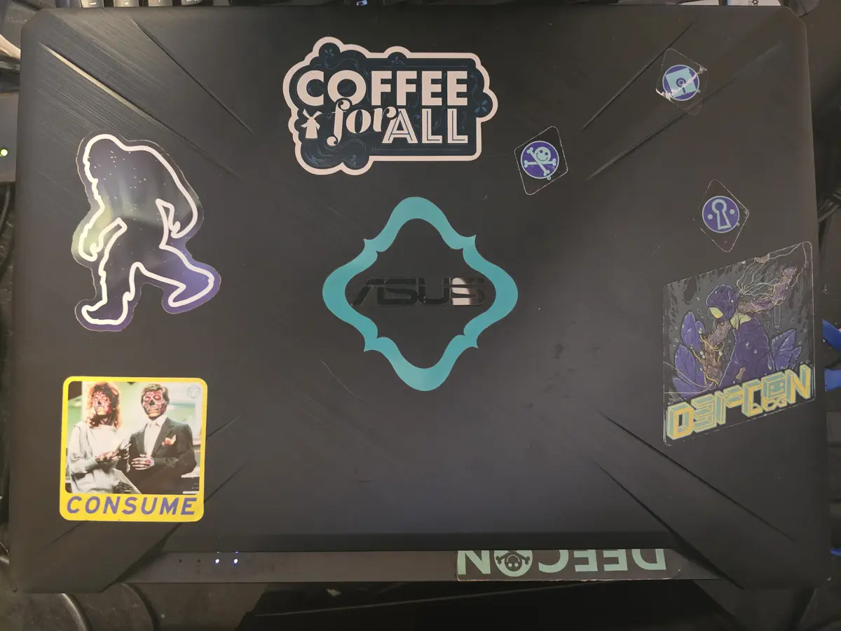 A black ASUS laptop lid covered in various stickers, including 'COFFEE for ALL', a Bigfoot silhouette, a 'CONSUME' sticker…