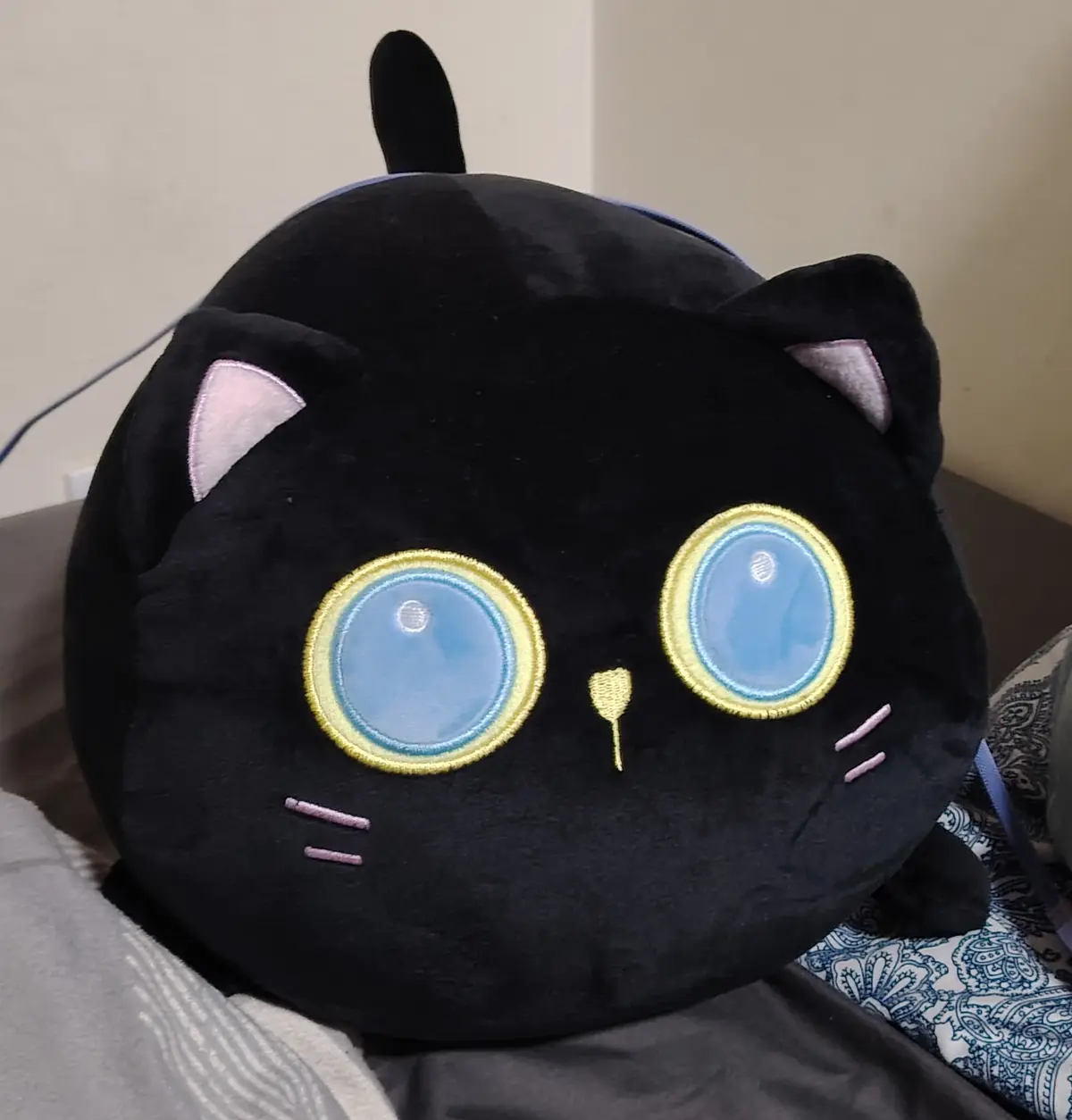 A close-up photo shows a large, round, black plush toy shaped like a cat's head, featuring oversized blue eyes with yellow…