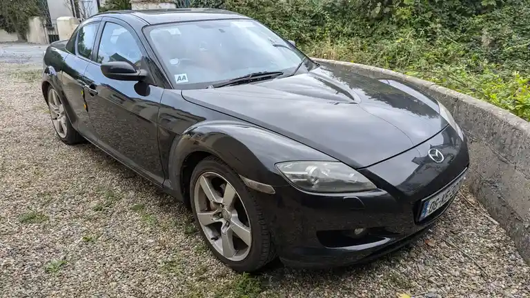 A black Mazda RX-8 sports car is parked on a gravel driveway next to a concrete curb and green foliage. The car has a glossy…